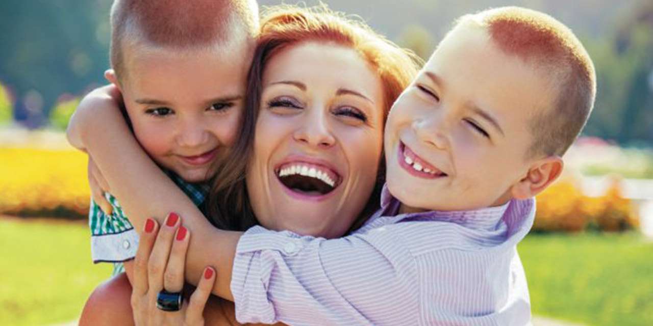 Una giovane mamma sorride abbracciata da due bambini in un parco