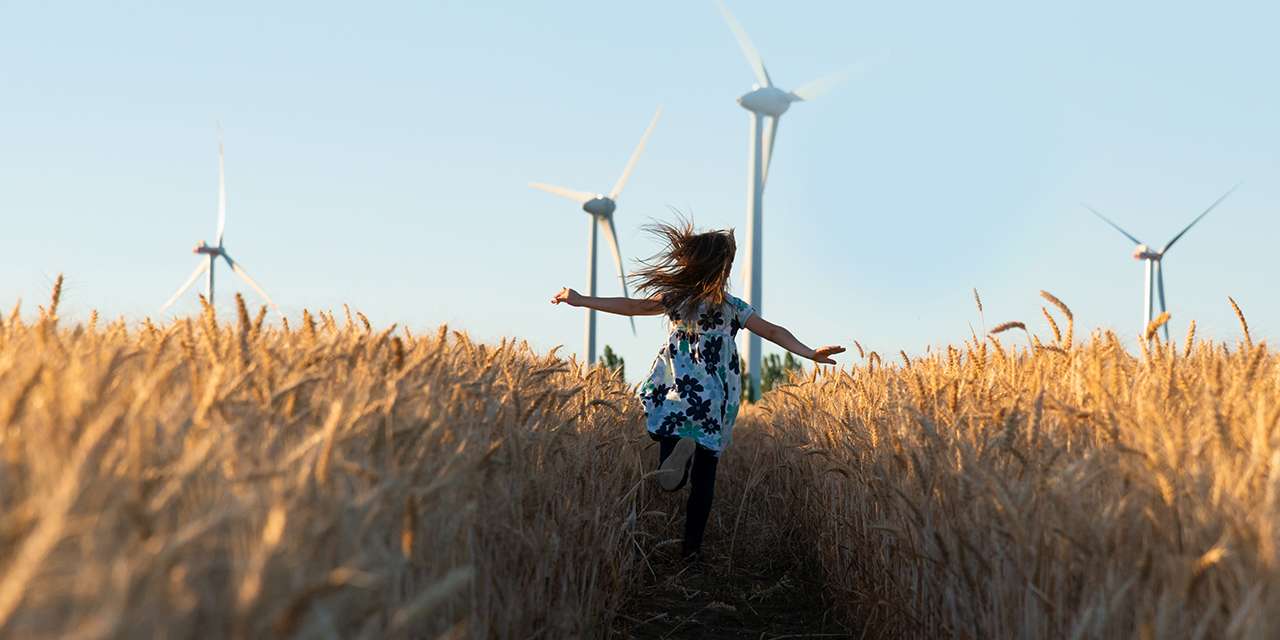Una bambina corre tra spighe di grano in un campo con pale eoliche: natura e fonti rinnovabili, un binomio per la sostenibilità