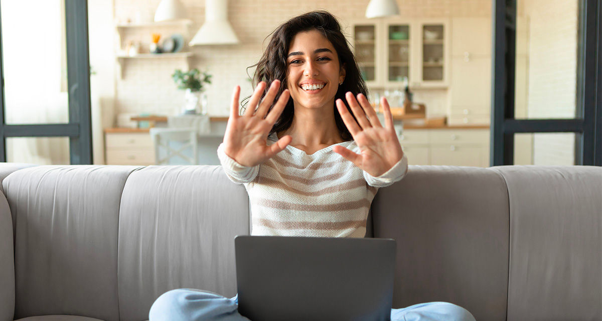 Una ragazza su un divano sorride davanti allo schermo di un portatile, mentre mostra i palmi delle mani