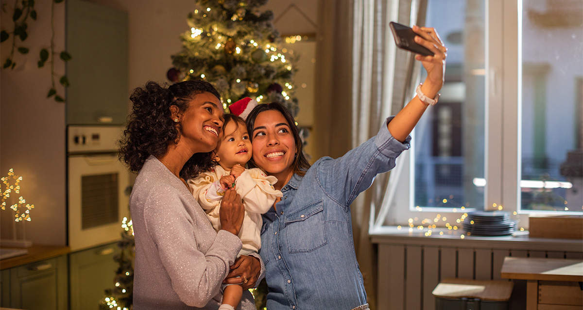 Due ragazze abbracciano e stringono sorridenti una bambina, mentre scattano un selfie di famiglia in una cucina decorata per Natale.