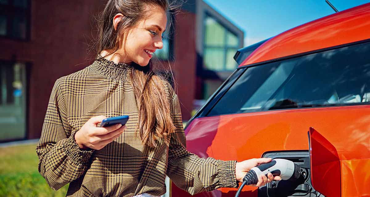 Una ragazza con lo smartphone inserisce la spina nella presa di ricarica dell’auto elettrica