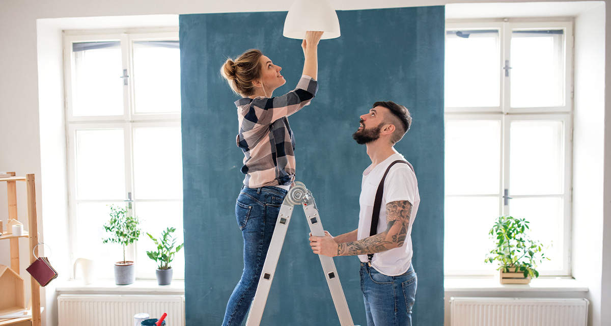 Una ragazza, su una scala sorretta da un ragazzo, avvita una lampadina al soffitto in un soggiorno con dei lavori di pittura in corso