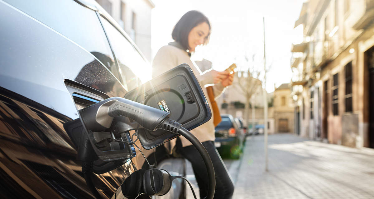 Una ragazza, appoggiata alla propria auto elettrica in ricarica, controlla lo smartphone